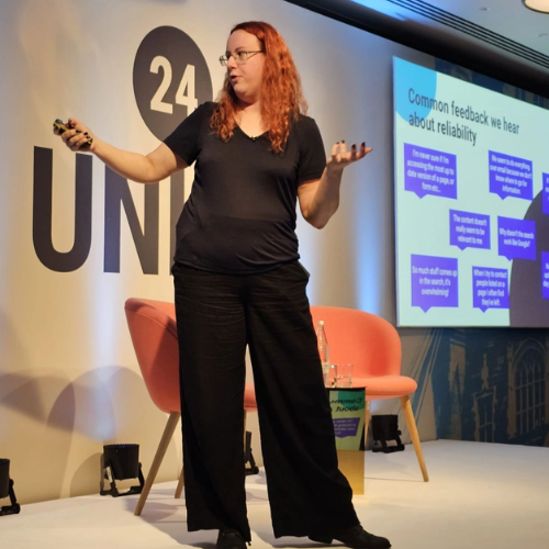 Speaker presenting on stage at Unite 24 conference with slides about intranet reliability feedback displayed on screen.