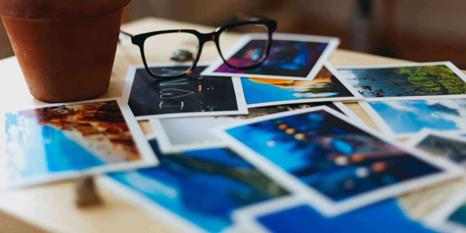 Close-up of printed photographs scattered on a wooden table beside a pair of glasses and a terracotta plant pot, evoking creativity, reflection, and storytelling.