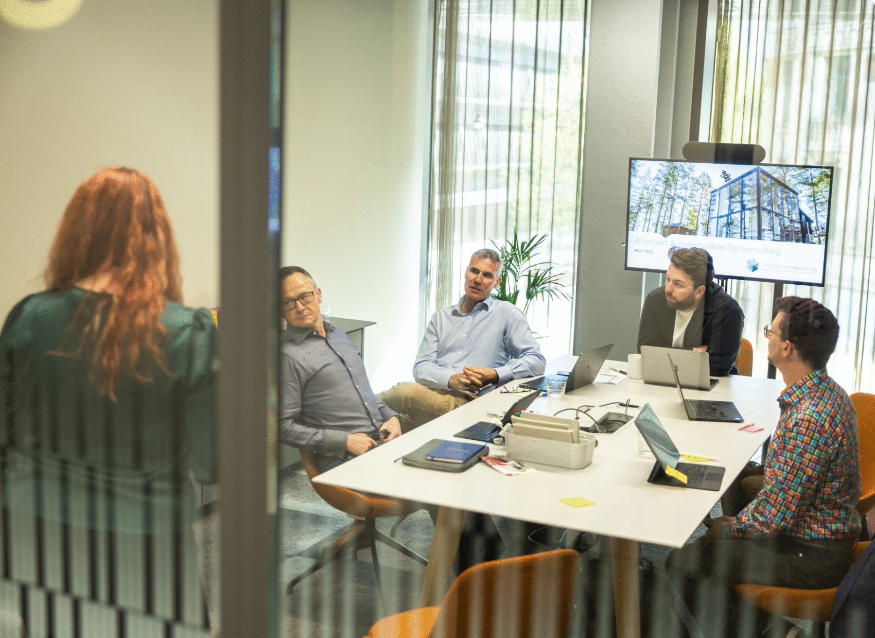 Business meeting in a modern office, with five people seated around a table and one standing, discussing a presentation on AI opportunities for consulting.