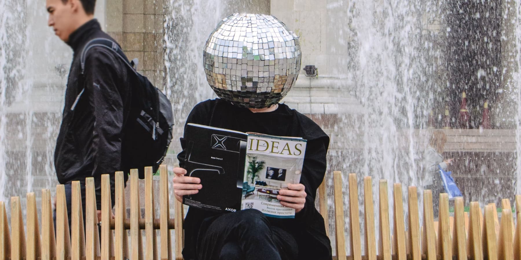 A person on a public bench with a disco / mirrored ball over their head, reading a magazine called 'Ideas'.