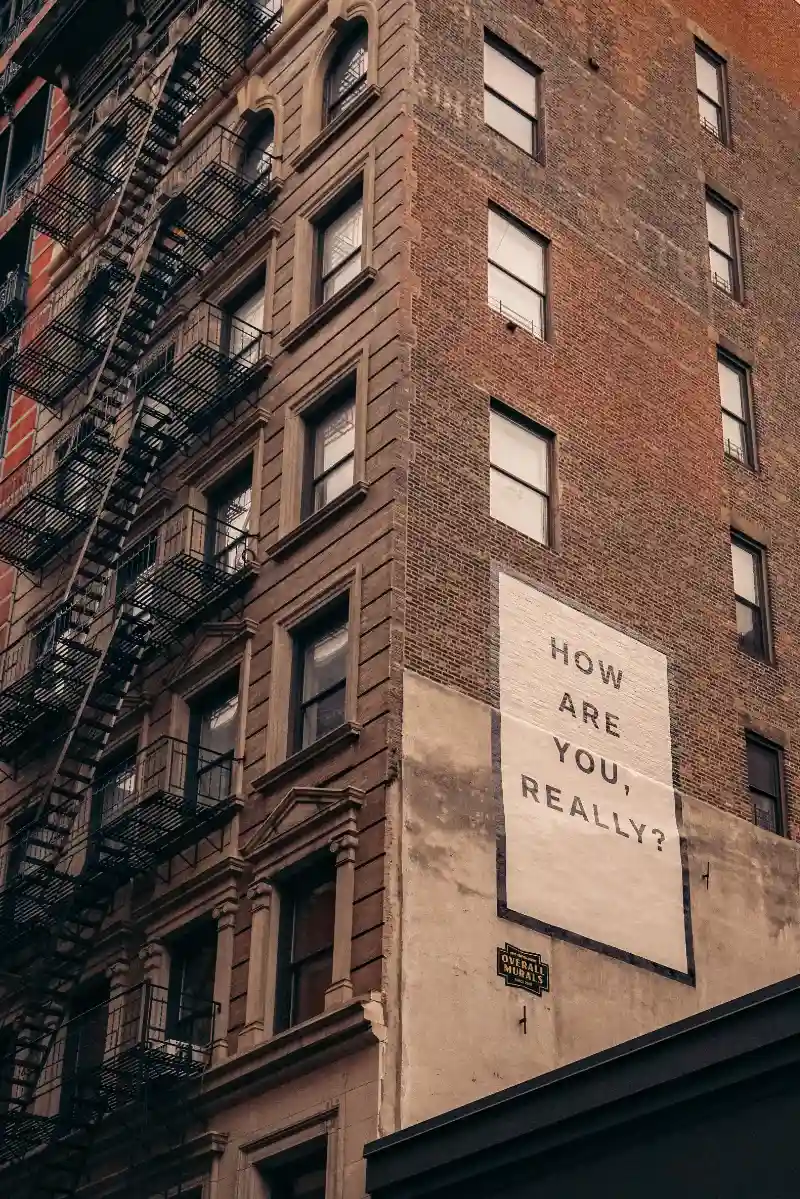 A brick building, fire escapes zig zagging up it. On the side, a simple poster; "How are you really?".