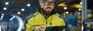 Man, glasses and hard hat, uses a tablet while wearing a high visability jacket in a warehouse.