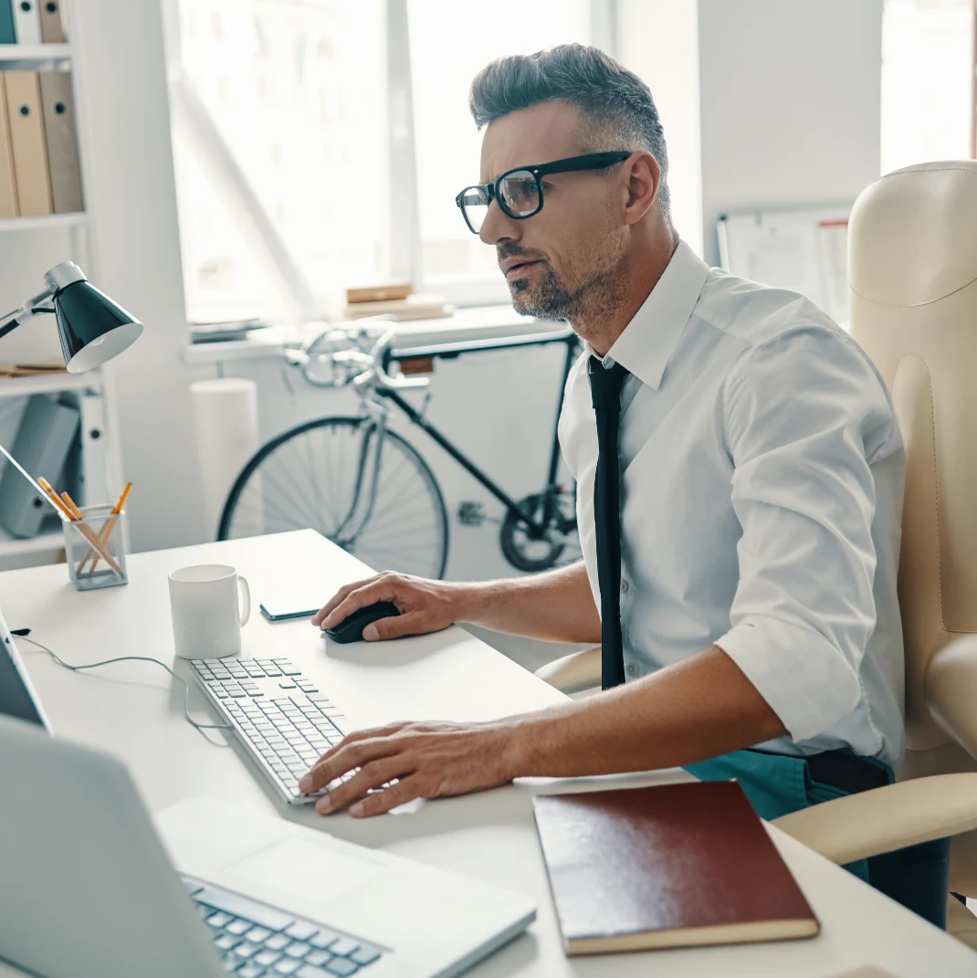 Man sat using his desktop computer with a laptop near by. White man in glasses, shirt and tie, might be an office but there's a bike in the background.